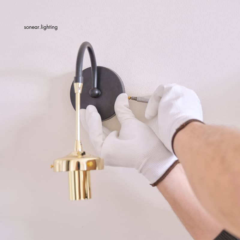 The process of installing a wall sconce with a black base and golden details, hands in white gloves tightening the fixture.
