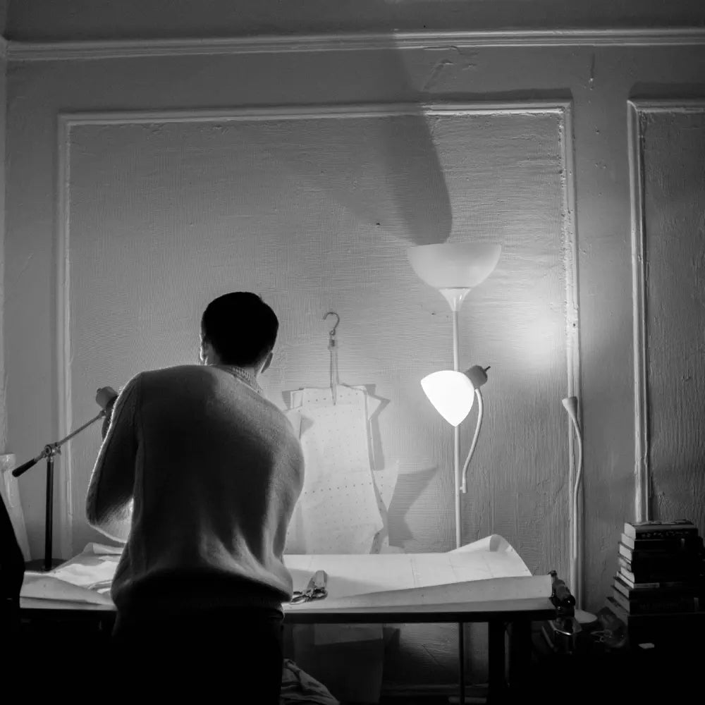 Černobílá fotografie designéra zády k fotoaparátu u stolu s papíry, nůžkami a šablonou, osvětleného dvěma stolními lampami s bílými stínidly – novinky v ruční výrobě LED osvětlení