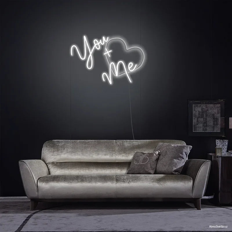 Bílý neonový nápis You + Me se srdcem, moderní světelná dekorace nad šedou pohovkou.