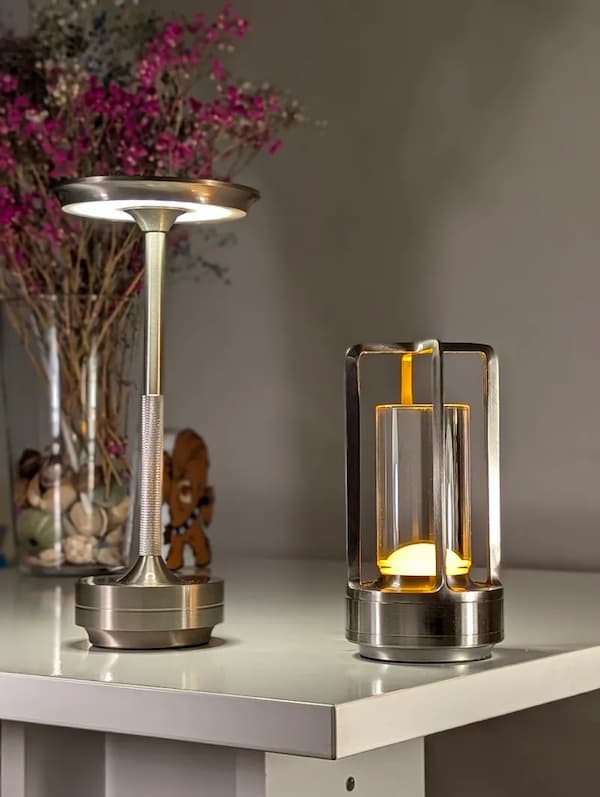 Stylové stolní lampy z kovu — moderní dobíjecí LED lampy s elegantním designem na bílém stole, v pozadí květiny ve váze.